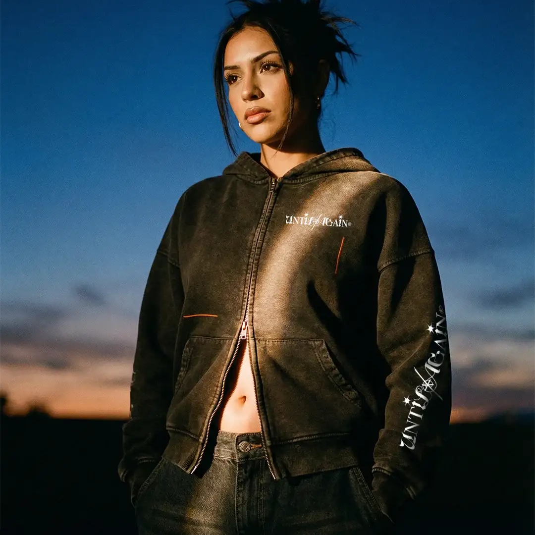 Woman in black streetwear hoodie with UNTIL/AGAIN logo at sunset, urban fashion style