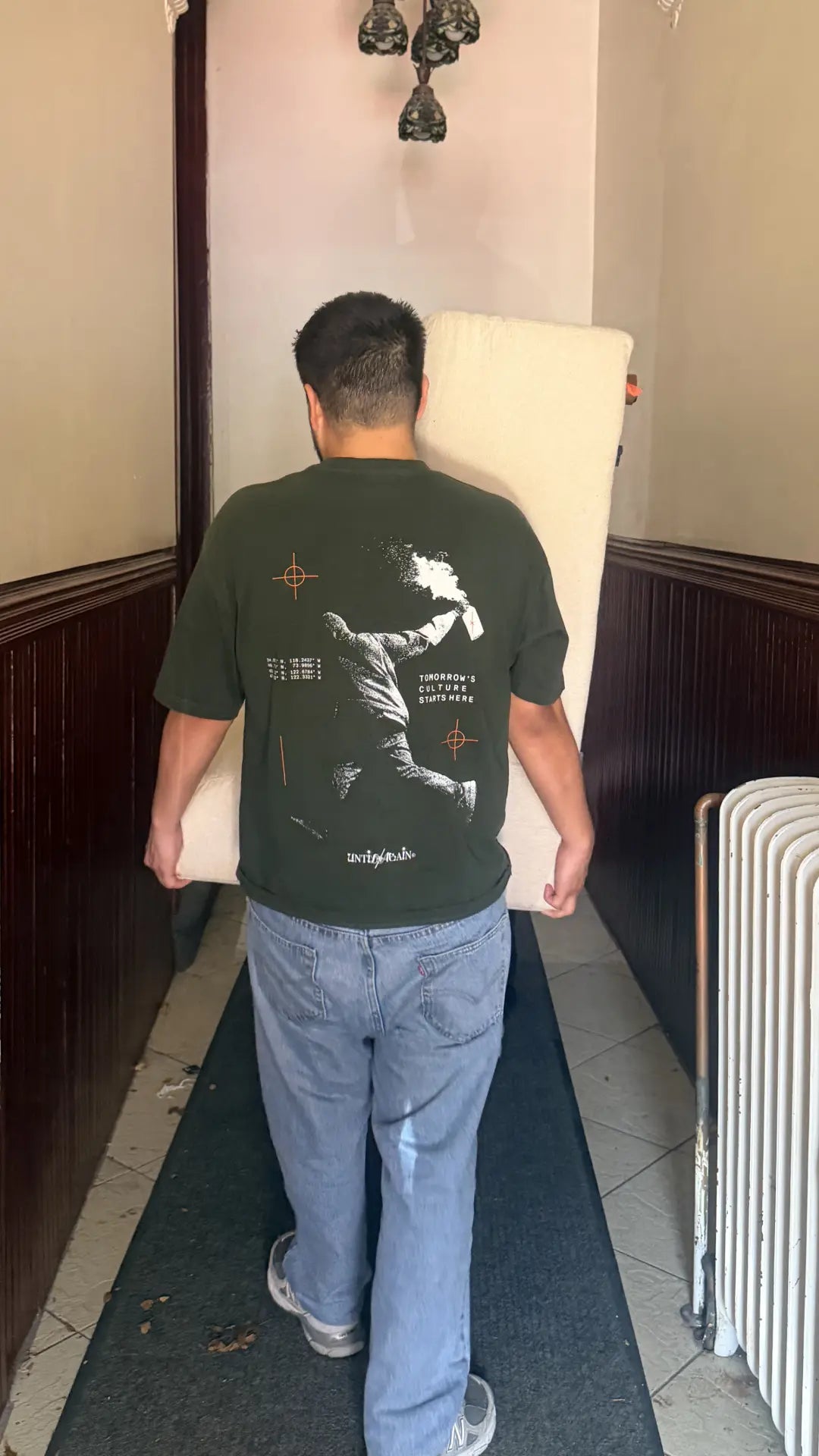 Man in UNTIL/AGAIN limited drop graphic tee carrying cushion a couch down the hallway of an older building in new york