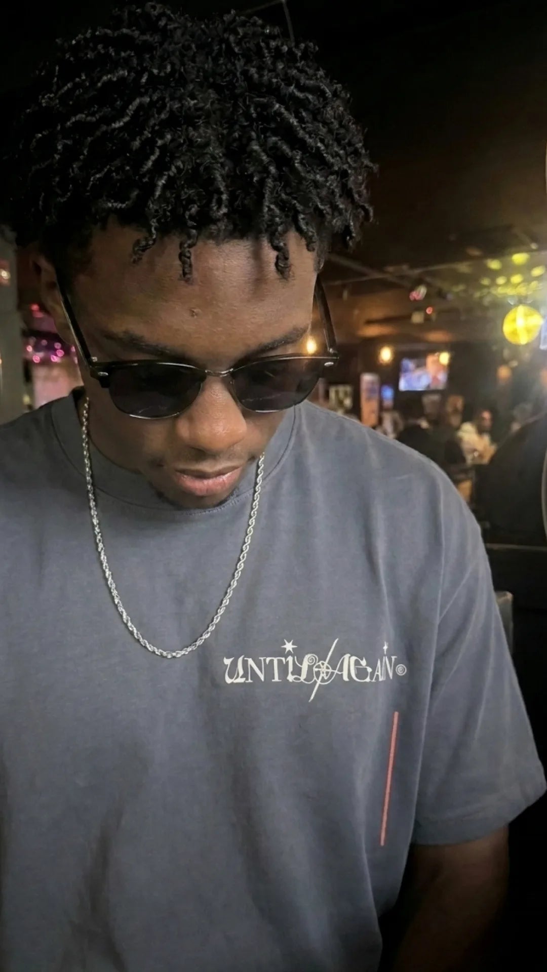 Man wearing sunglasses and a limited drop UNTIL/AGAIN t-shirt in a dimly lit setting
