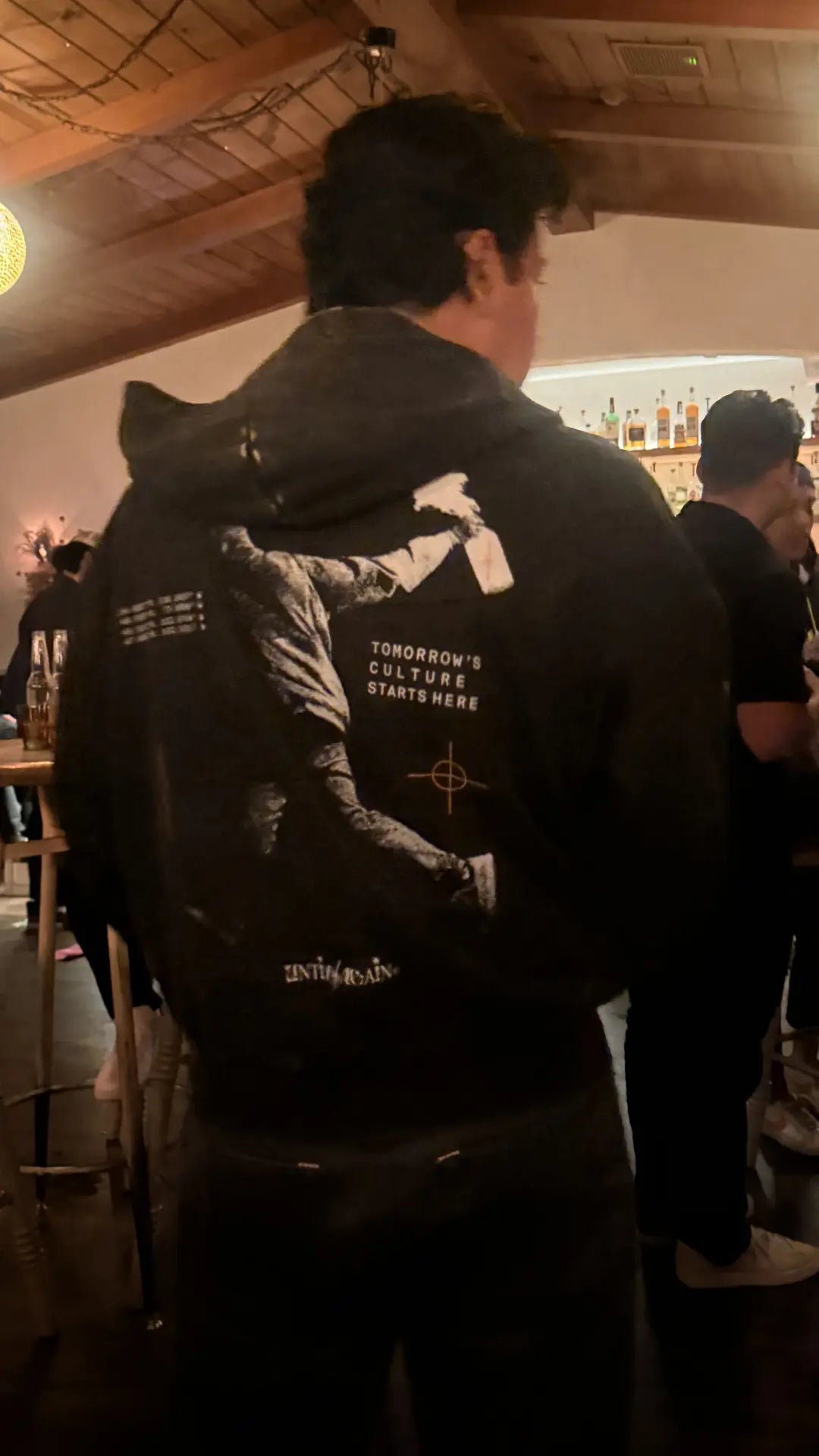 Person wearing UNTIL/AGAIN limited drop hoodie with bold graphic and text at a social event