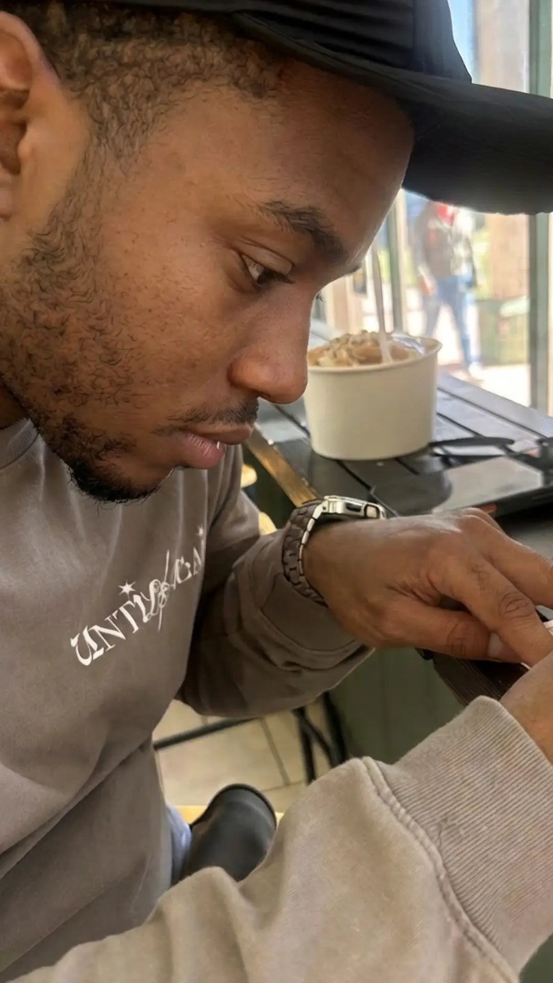 Man in UNTIL/AGAIN limited drop sweatshirt focused at a table, casual cafe setting