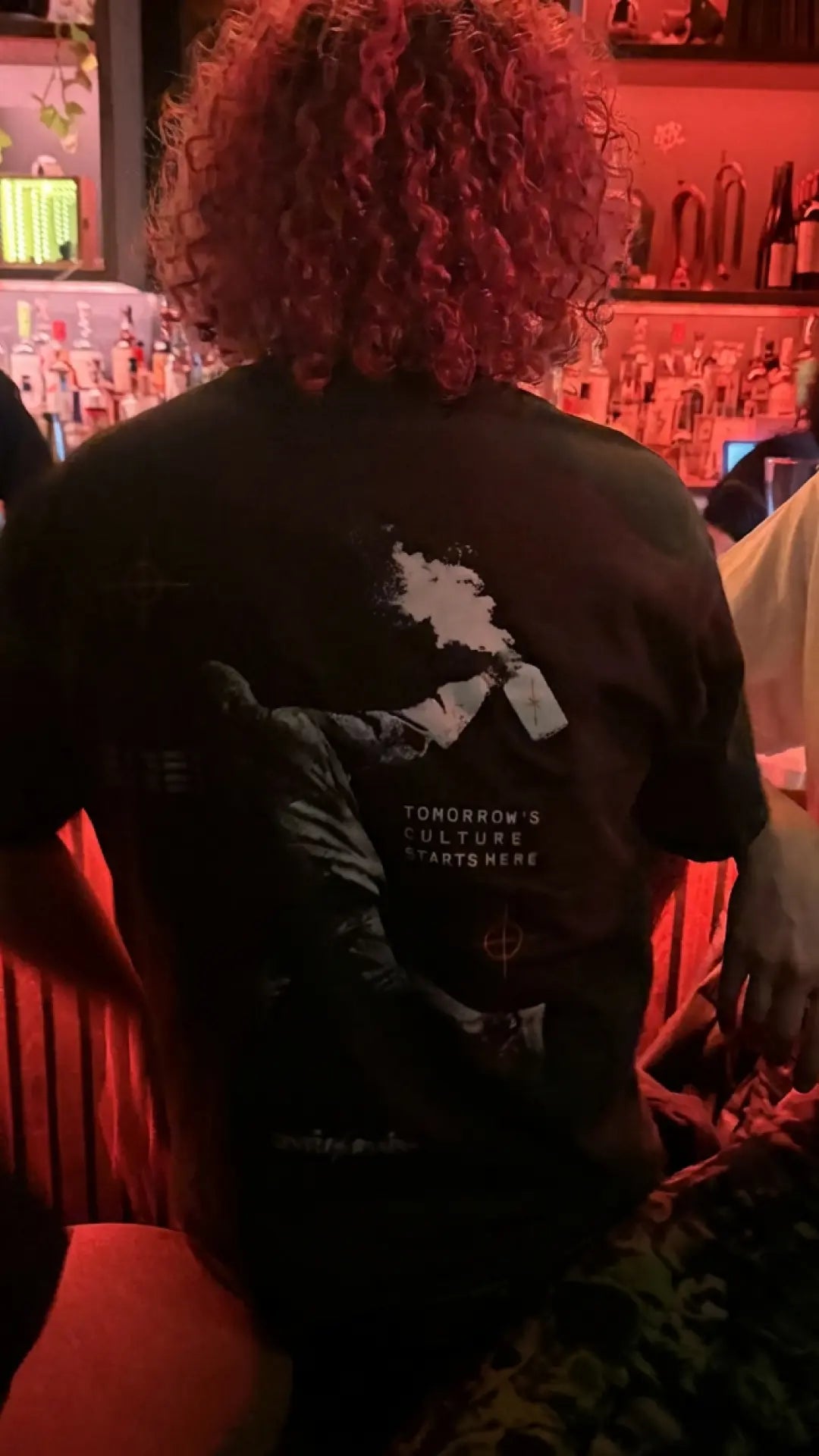 Person in a bar wearing a black UNTIL/AGAIN limited drop tee with bold back graphic.
