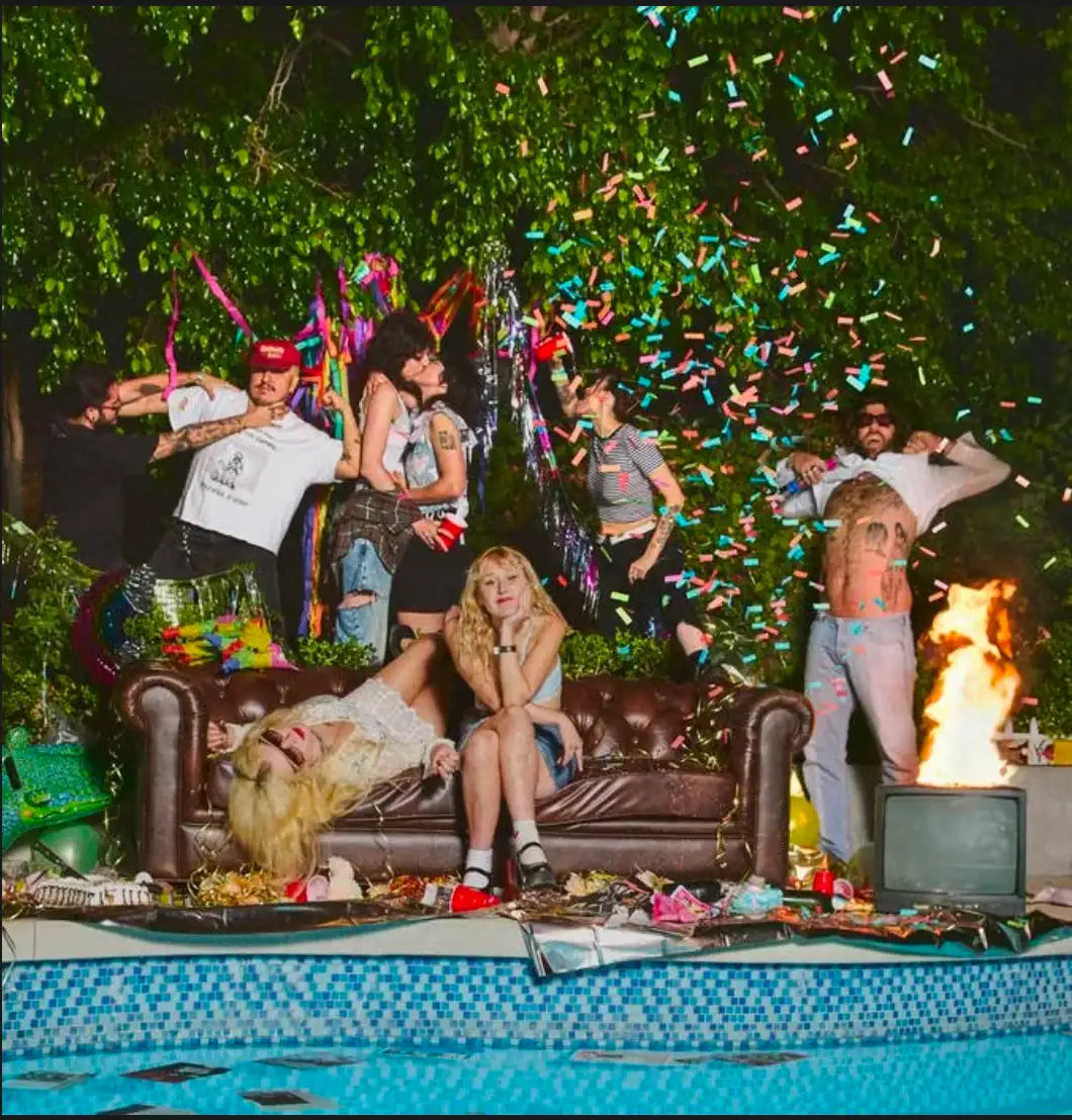 Group of young adults at an outdoor pool party with confetti, vintage couch, fire, and retro TV.