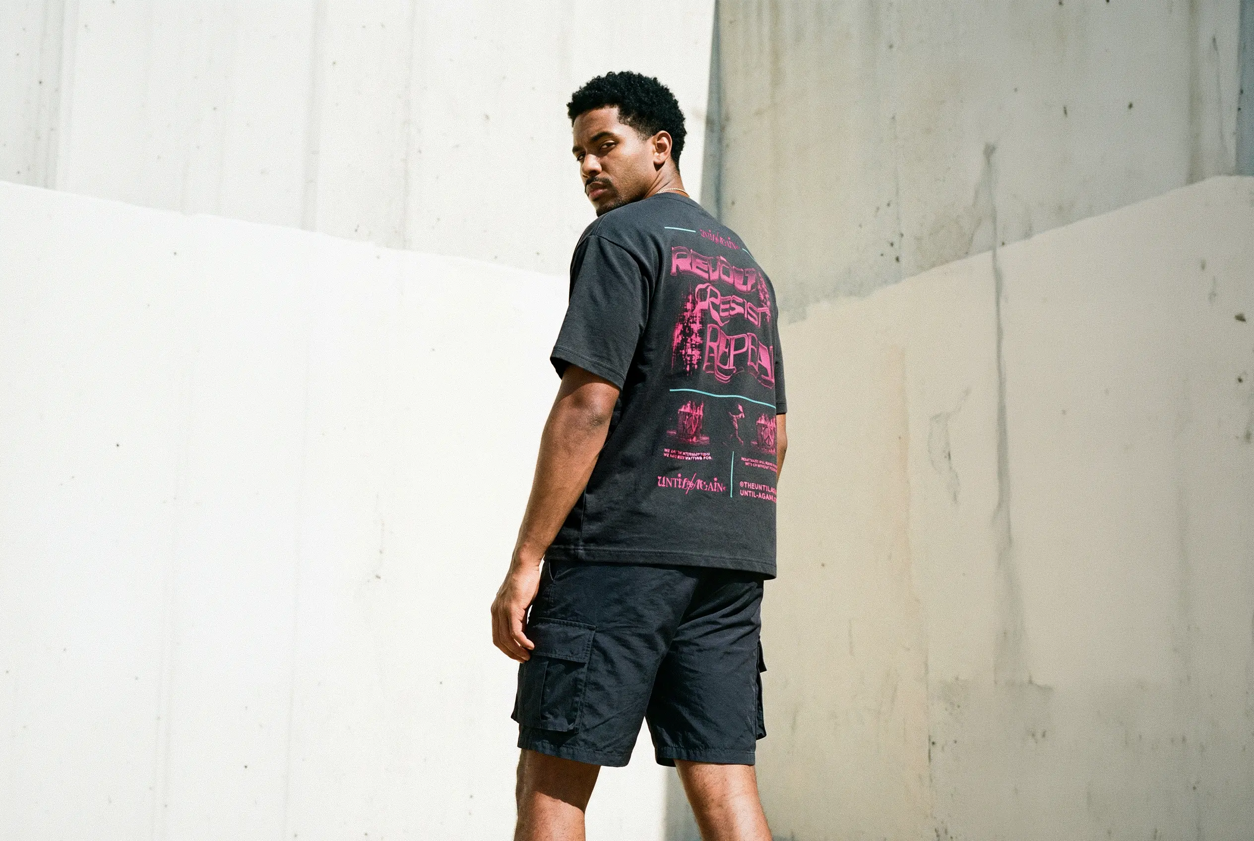 Man wearing a black t-shirt with pink graphics and dark shorts against a concrete wall.