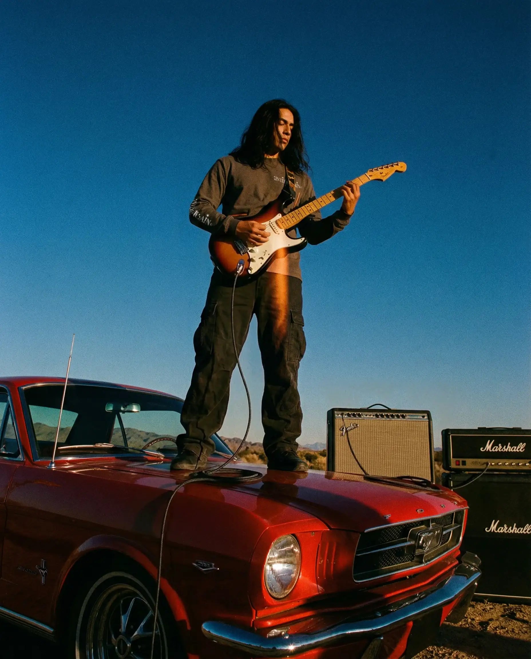 Man in streetwear plays electric guitar on red vintage Mustang with amplifiers outdoors Tomorrow’s Culture Long Sleeve charcoal front clean minimal streetwear