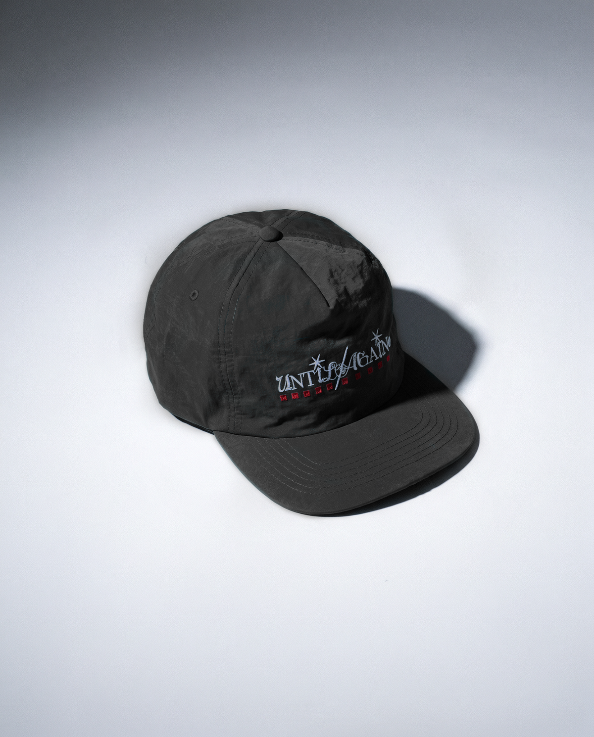 A black nylon baseball cap with a curved brim, featuring a white embroidered logo that reads ''UNTIL AGAIN' with red squares below it, set against a light gray gradient background.