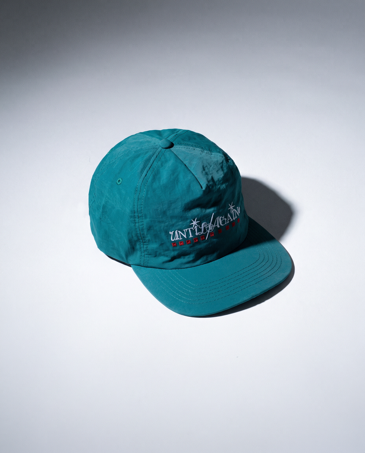A teal baseball cap with embroidered white and red text reading 'UNTIL AGAIN' on the front, photographed on a clean white studio background with a soft shadow.