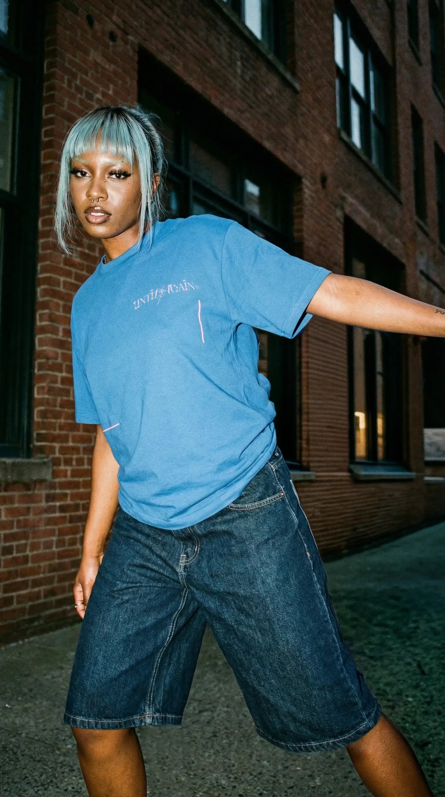 Model wearing blue streetwear t-shirt and denim shorts in urban brick alley setting